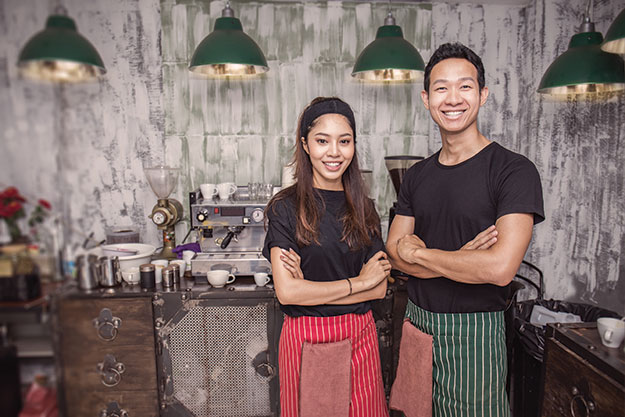 Two colleagues standing in the kitchen with arms crossed, smiling at the camera wearing aprons.